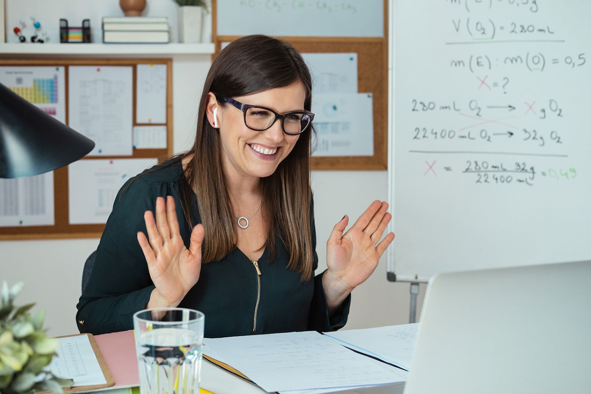 An educator looking at laptop screen and waving with hands, greeting e-learning students to online lesson.