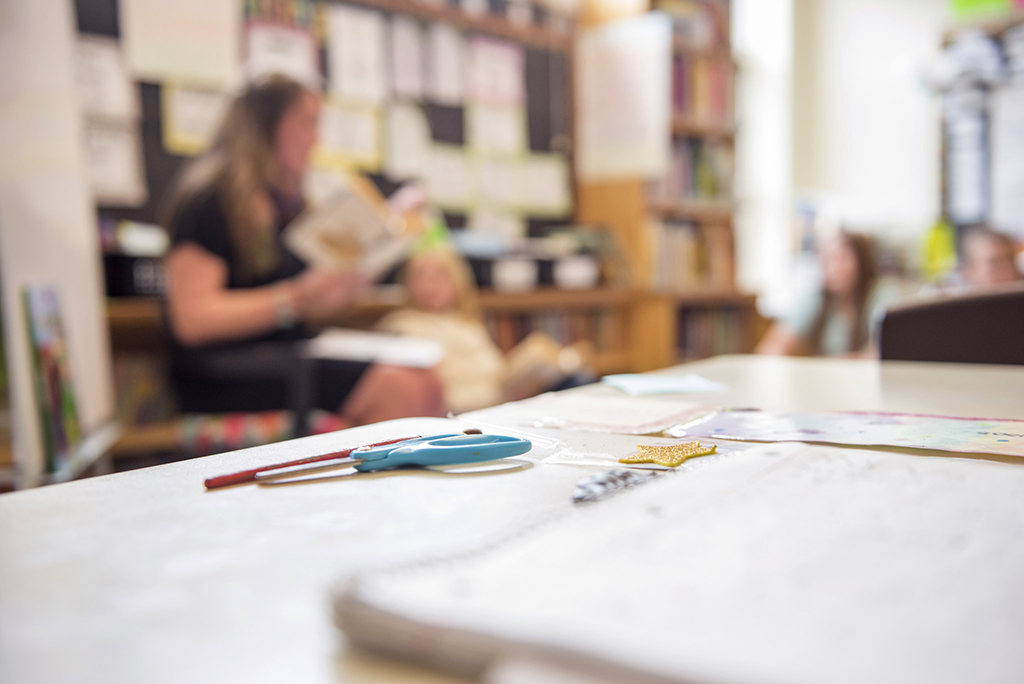 A close-up photo of a notepad and pen on a table, with a teacher instructing students in the far background