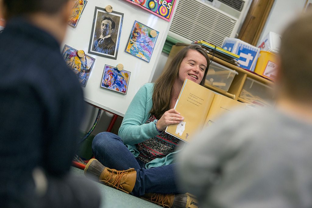 A photo of a teacher holding a book, pages opened facing the children seated before her.