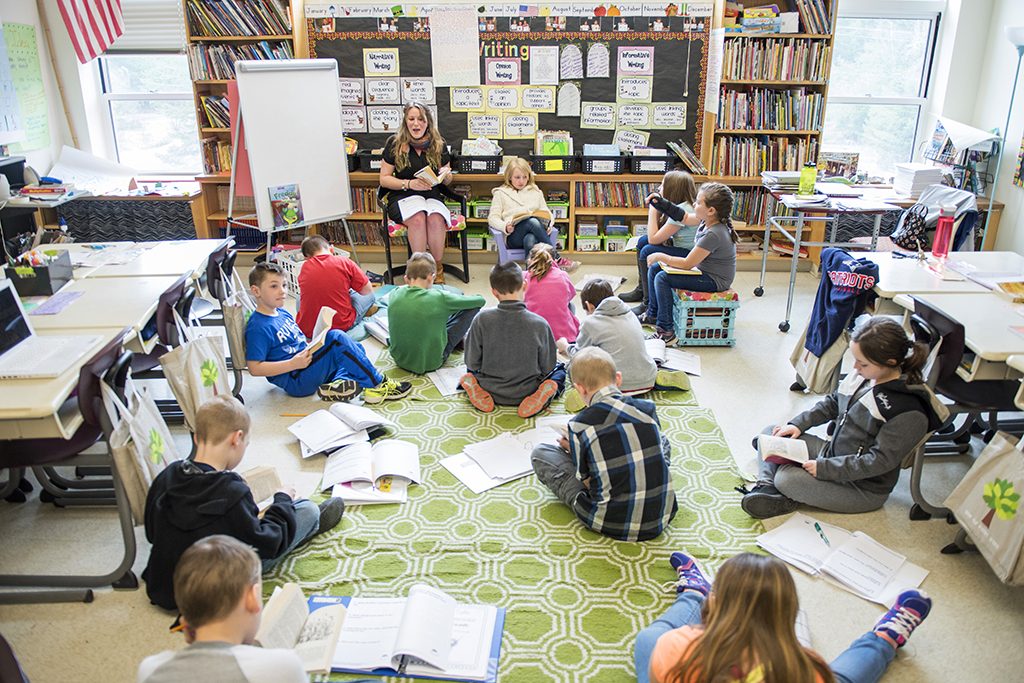 A grade school classroom setting, with many students seated on the floor, papers laid about them.