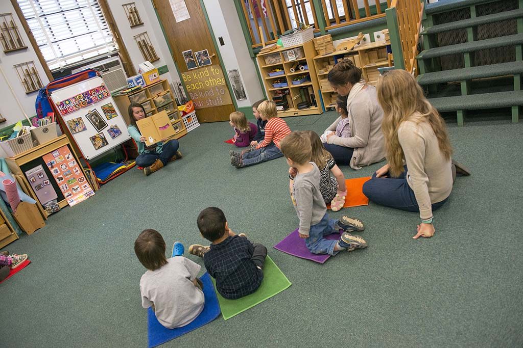 A photo of children seated in a semicircle on the floor of a classroom while student teachers interact with them.