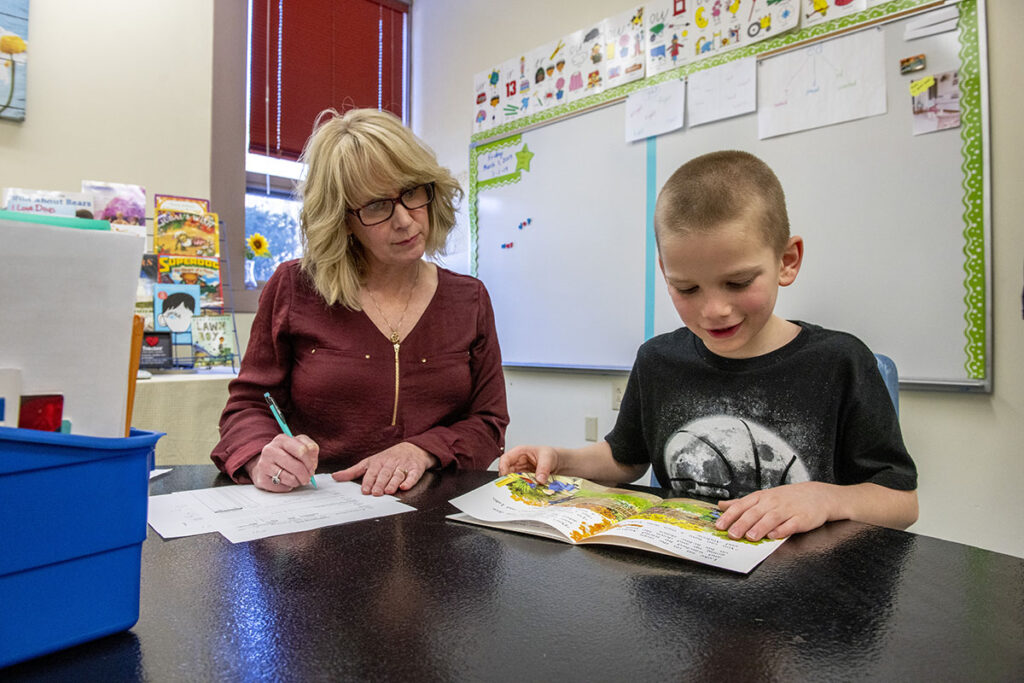 A photo of a teacher with a child at a classroom table, together looking at an open book.