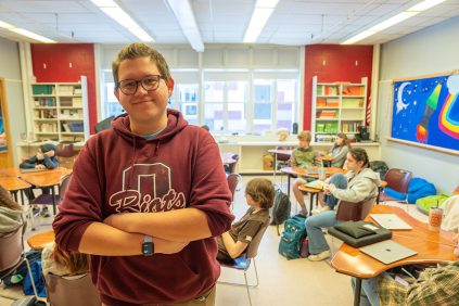 A portrait of University of Maine alumni Jarod Webb posing in front of his classroom at Orono High School.