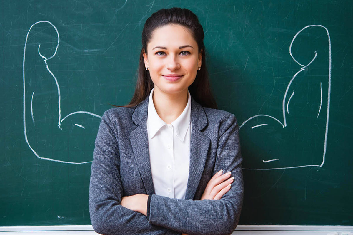 A female teacher with her arms folded stands in front of a chalkboard with flexing biceps drawn behind her.