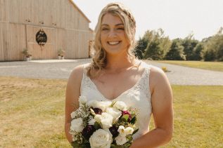 Carly Frost (McDonald) poses in her wedding dress with a bouquet of flowers in an outdoor setting with a barn in the background.