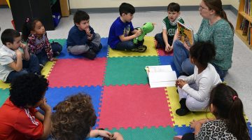 A teacher sits on a mat in a circle reading to a group of young children.