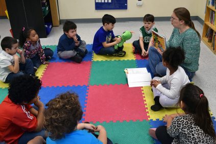 A teacher sits on a mat in a circle reading to a group of young children.