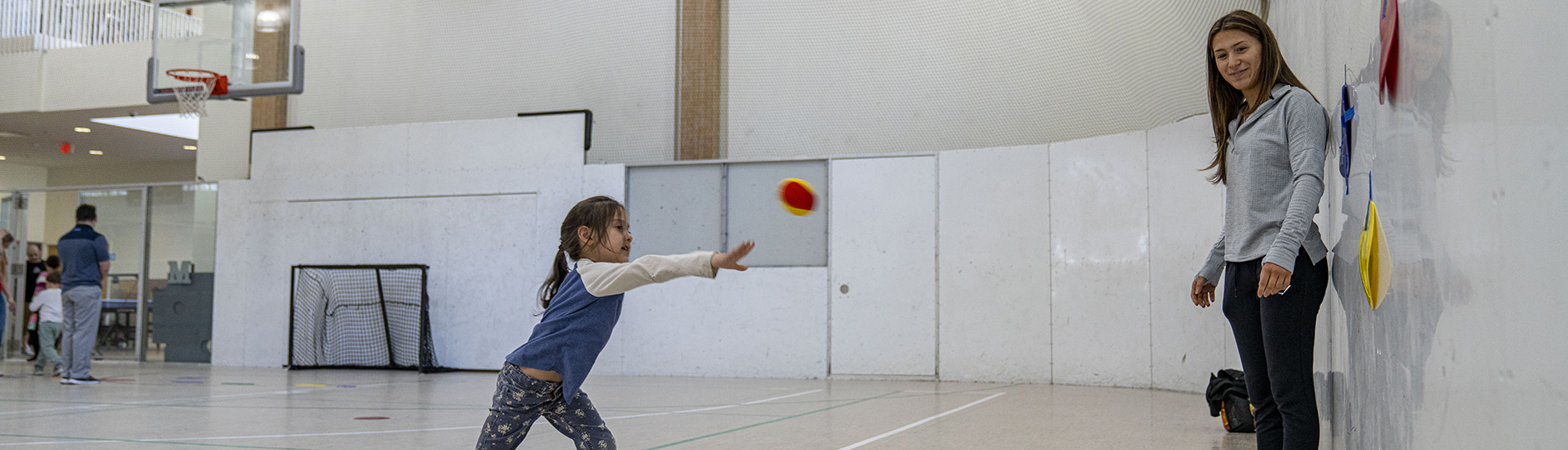 A photo of a young girl working with a University of Maine student on throwing a ball in a gym.