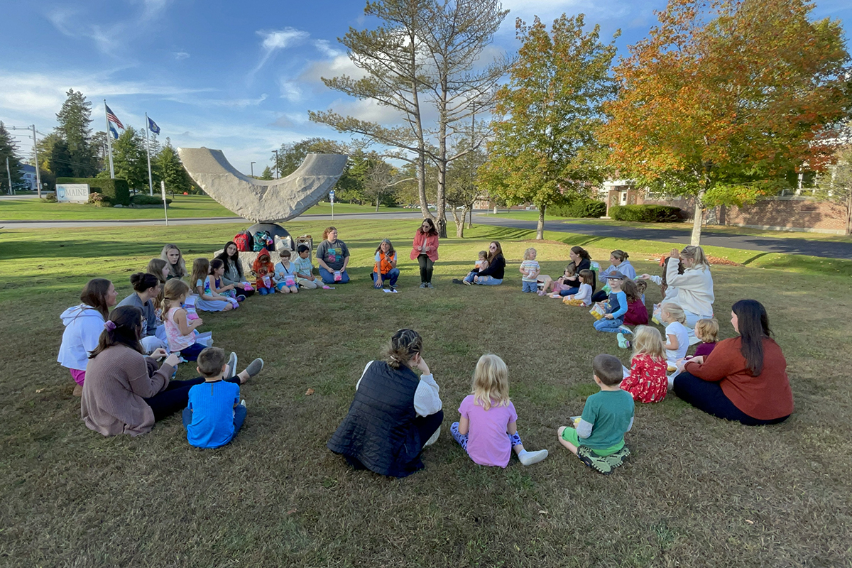 A group of children and University of Maine students sitting in a circle on a lawn.