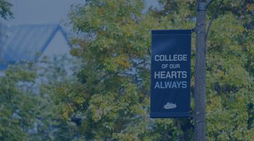 A photo with a blue color overlay showing a flag on the University of Maine campus with the text "College of Our Hearts Always."