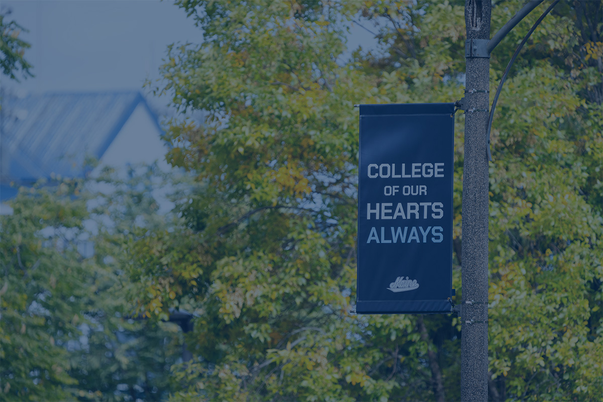 A photo with a blue color overlay showing a flag on the University of Maine campus with the text "College of Our Hearts Always."