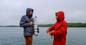 Two people stand on a dock holding sampling materials while wearing raincoats.