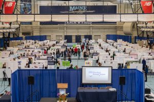 Elevated perspective of the presentation floor at the UMaine Field House during the Student Symposium. A small stage space with a projector screen and a lectern can be seen in the foreground, separated from the rest of the space by blue curtains. Behind the stage are hundreds of large posters displayed on easels in rows.