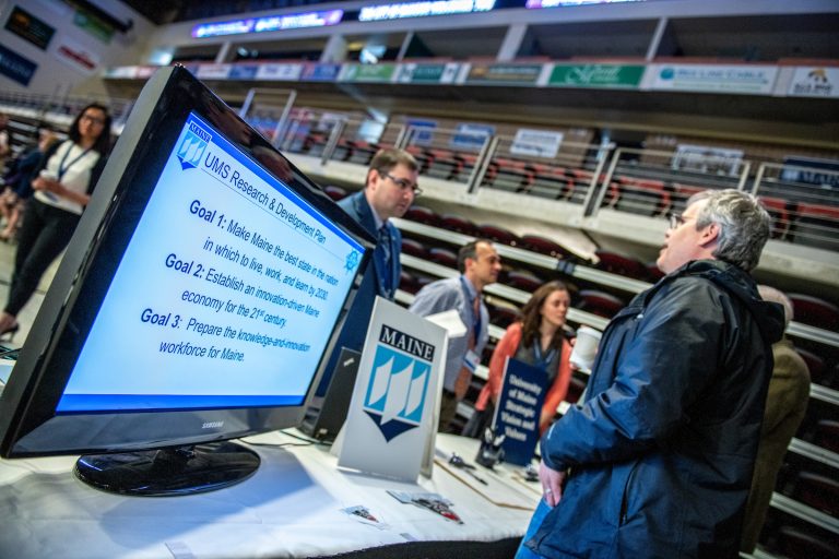 An image from the UMaine Student Symposium in 2019. A display table is seen in the foreground where bleacher seating for Bangor Maine's Cross Insurance Center arena has been pushed back. On the table are a few signs with UMaine Branding as well as a digital display screen on the far left. The screen shows a slide titled "UMS Research & Development Plan" and lists three goals. Three people with blue lanyards and nametags stand behind the table, speaking to visitors.