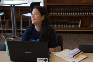 Melissa Kimble woring on a computer at the Univeristy of Maine Library. She is sitting at table with her laptop with books on a shelf behind her. 