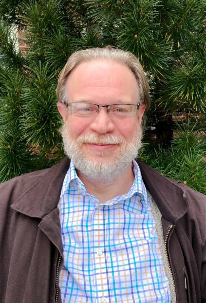 Christian Spindler stands in front of a conifer wearing a brown jacket