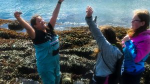 Three women stand in tide pool, two with their arms raised in celebration.