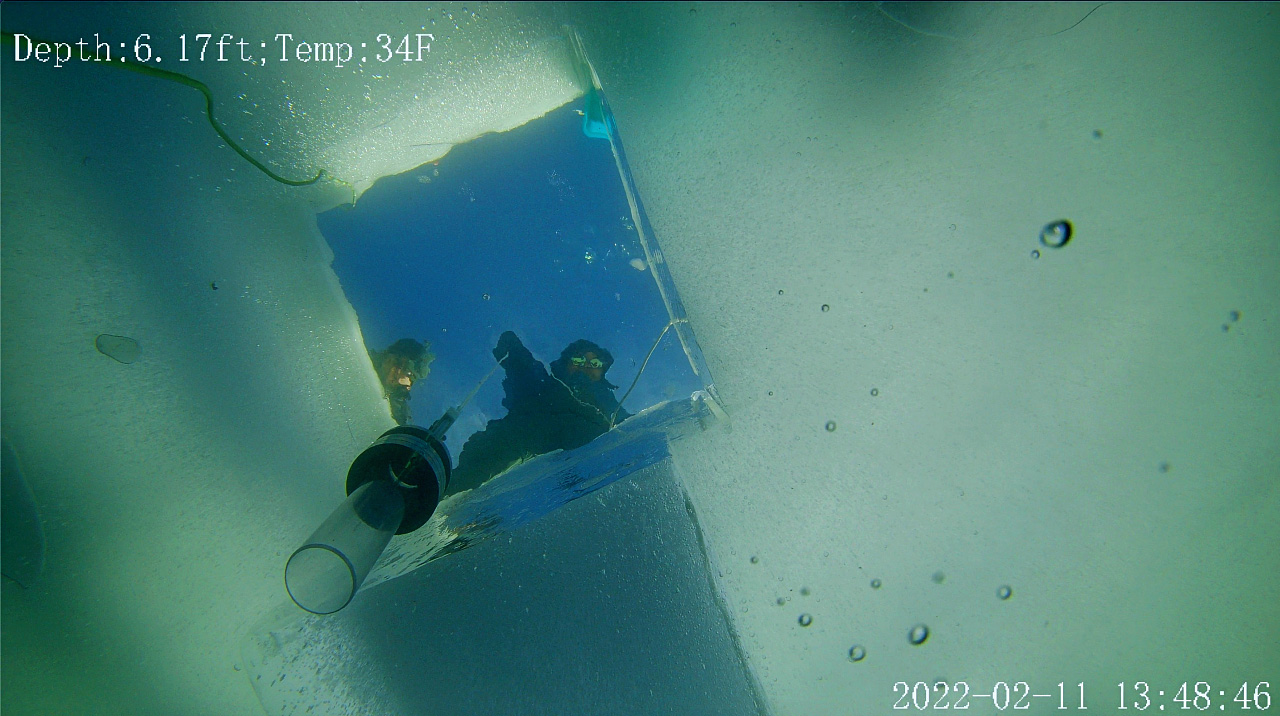 Two individuals stand above hole in the ice as a sampling tool is pulled up.