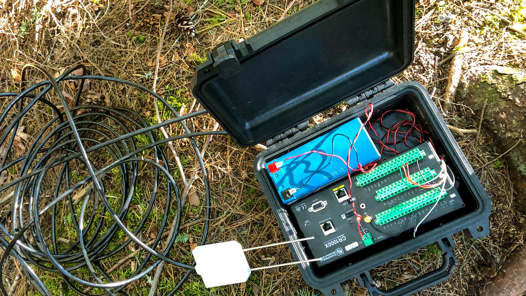 Black carrying case with electronics sits on the ground with cables extending from it.