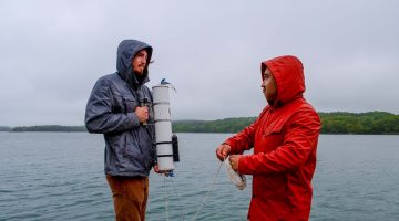 Two people stand on a dock holding sampling materials while wearing raincoats.