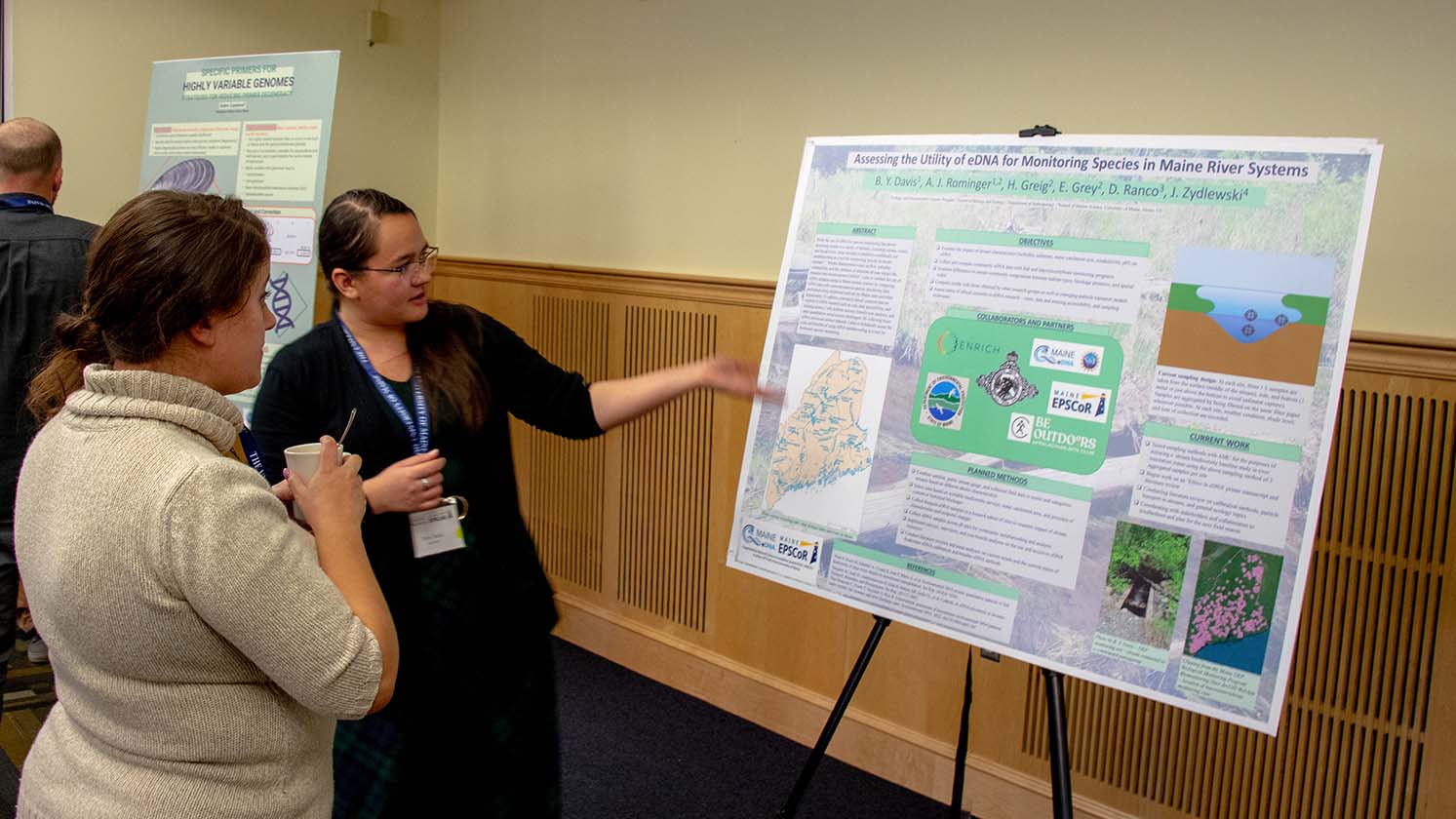 Two people stand indoors as one presents a poster to another.