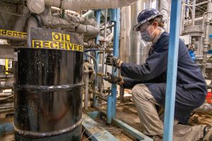 Researcher wearing hard hat crouches next to large barrel with text that says "Oil Reciever"