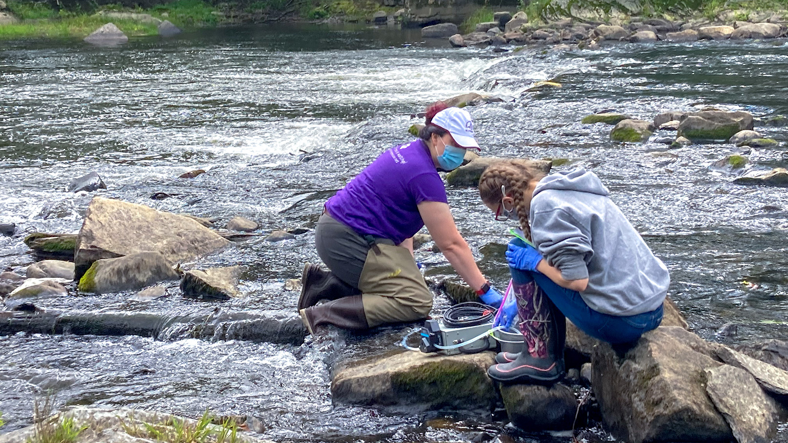 Two people kneel in river collecting water sample.