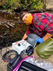 Masked person in flannel and waders kneels on rock taking water sample. 