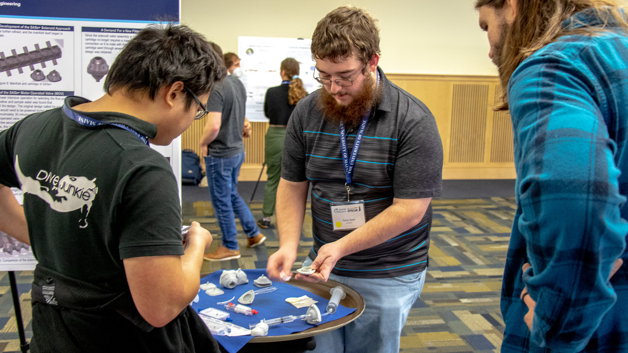 Two individuals gather around Aaron Snow as he shows his 3D printed parts at research symposium.