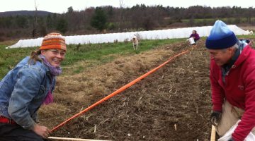 A student smiles at the camera as she helps mark a plot in a farm field. Another person is also setting lines for the plot in the foreground. Another person, an high tunnel, and a forest is in the background.