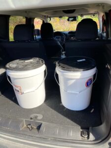 two white, lidded 5 gallon buckets in the back of a car.