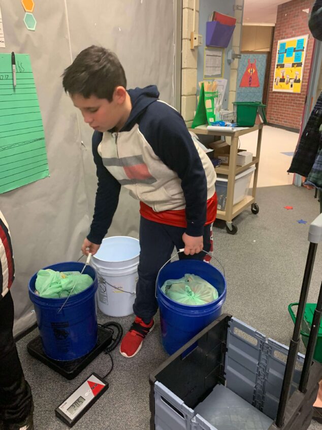 picture of a fifth grade student from the Gerald E. Talbot Community School in Portland, Maine. Student is weighing a bucket of food waste.