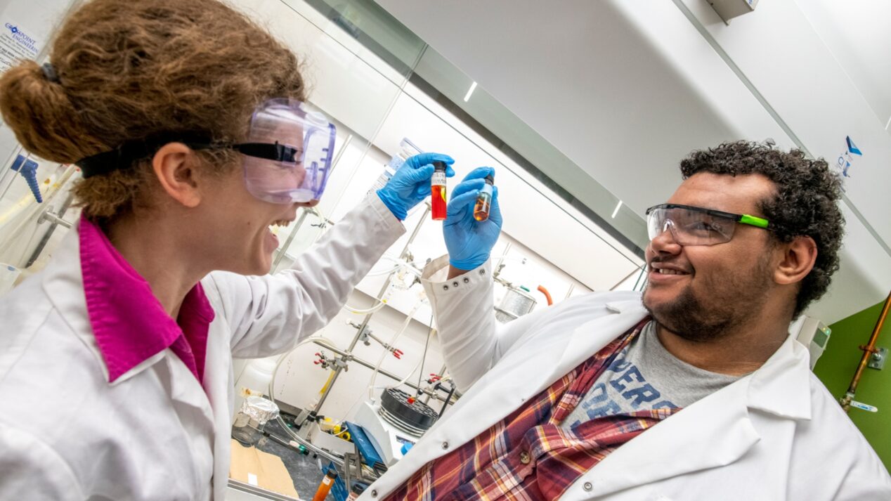 two lab students smiling at each other while holding vials
