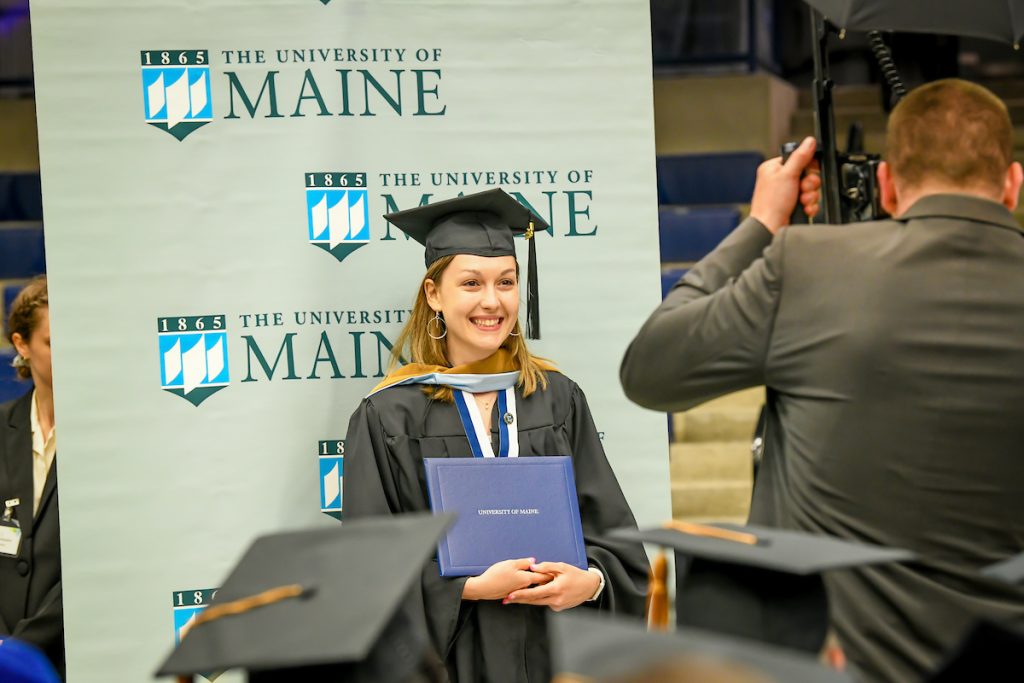 Graduate getting their photo taken with their diploma against a UMaine themed background