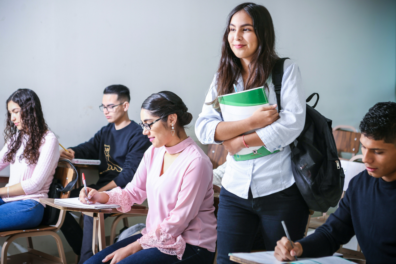 Student standing and holding books in the middle of a classroom of students sitting at desks