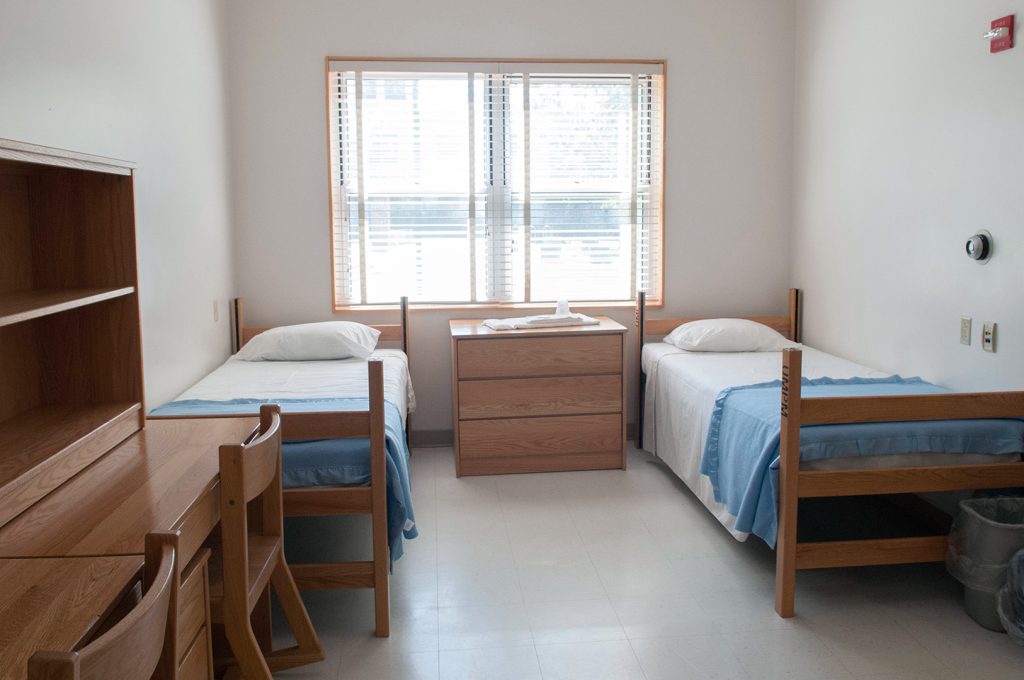 Dormitory room containing two single beds, a dresser under a window, and a desk.
