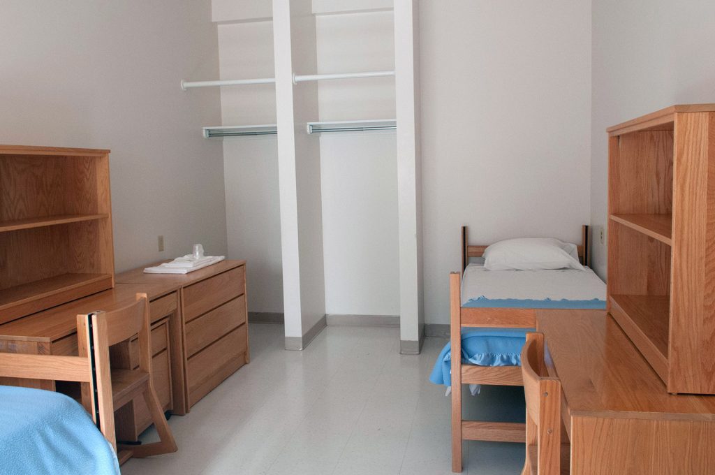 Dormitory room with wooden desks, chairs, a single bed, and open closet spaces.
