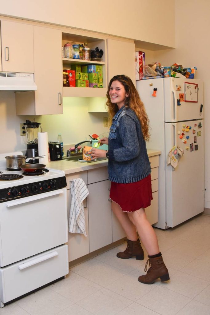 A student standing by a kitchen sink near a white stove and refrigerator.