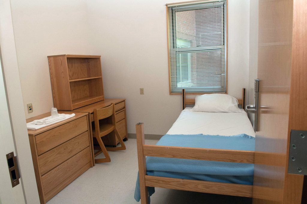 Dorm room with a single bed, wooden desk, chair, dresser, and window.