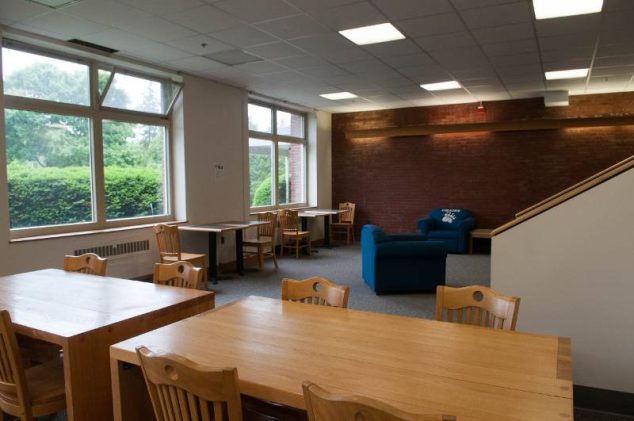 Study lounge with wooden tables, chairs, windows, and blue armchairs near a brick wall.
