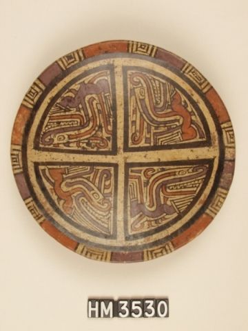 Polychrome ceramic plate Decorated in quadrants, each depicts a crested, curly-nosed iguana [see alternate description below], identified as such particularly by the horizontal eye slit and clawed limbs. Distinctive eye stripes indicate the theme of boa constrictor, frequently paired with the iguana. The rim pattern suggests the oval-rectangle sequence of markings on boa constrictors.