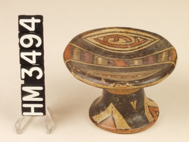 Cocle miniature polychrome ceramic pedestal plate. Pica Pica variety. This is miniature pedestal plate may have been used to hold a powdered snuff and probably represents a mushroom with a flat cap. The design on the top of the plate features a central bar as a serpent with boa ovals and rectangles represented by alternating red and cream bands. Adjacent hemispheres each contain a "Y" element with a claw or a curly crested "Y" element head and tail. In either case, an animal kenning is encased within boa ovals.