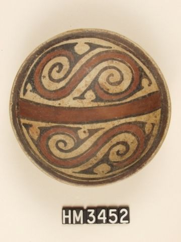 Cocle polychrome ceramic dish. The curvilinear red band in each of the hemispheres represents the body of a serpent, as does the central bar, also red. Black Y-elements depicting, in profile, heads of pointy-nosed animals with curly head crests are appended to each end of the serpent bodies. Additional Y-elements may indicate the location of limbs. The overall design expresses the fundamental theme of duality and the relatedness (the serpent body) that connects separate entities (the animals).