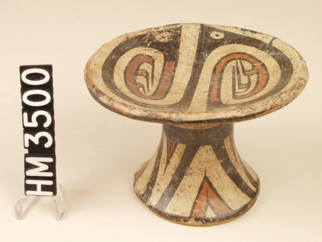 Cocle miniature polychrome ceramic pedestal plate. Macaracas type, Pica Pica variety. 
The basic design consists of four "Y" elements composing two connected hemispheres, suggesting a single serpentine body line in purple. Each end of the line is portrayed as a red "Y" element with the long end of the element depicted as either a claw or a curly head crest in profile. Color contrast suggests that the claw or crest represents the presence of a creature other than the serpent. The presence of eyes in the gaps of of the "Y" elements forming the serpent body line suggests that each hemisphere is composed of a double-headed serpent, that becomes a single double-headed creature of a different type.