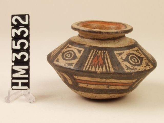 Miniature polychrome angled ceramic jar. Globular bottom, angled sides, narrow neck, and flared out-turned mouth. Mouth painted red with black border. Black line around rim and neck. Top half of body painted black with rectangles left buff with black circle, triangle, and linear designs inside. On opposite sides there are rectangles with red zig-zag patterns inside.