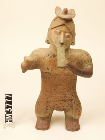 Ceramic standing figure with headdress playing reed flute with left hand and holding rattle in right hand.