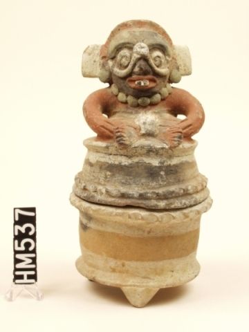 Covered ceramic container. Body supported on three small feet. Cover made with a masked figure sitting cross-legged on top.