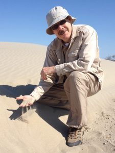 Image of a caucasian man in field clothes kneeling in sand.