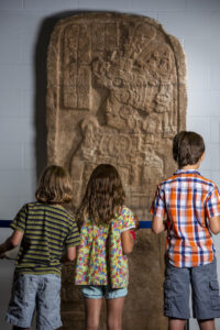 Three children stand in front of a Maya stele.