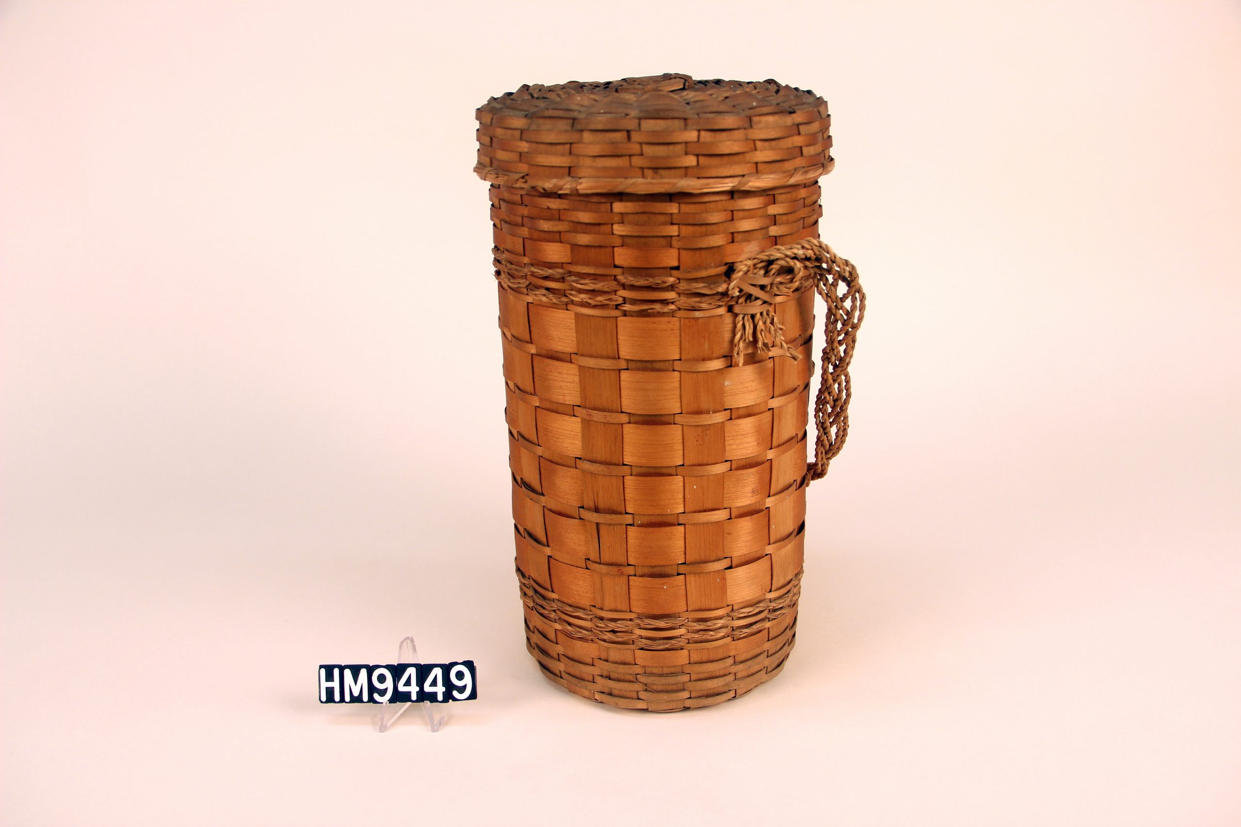 Image of a tall, cylindrical basket with cover and braided strap.